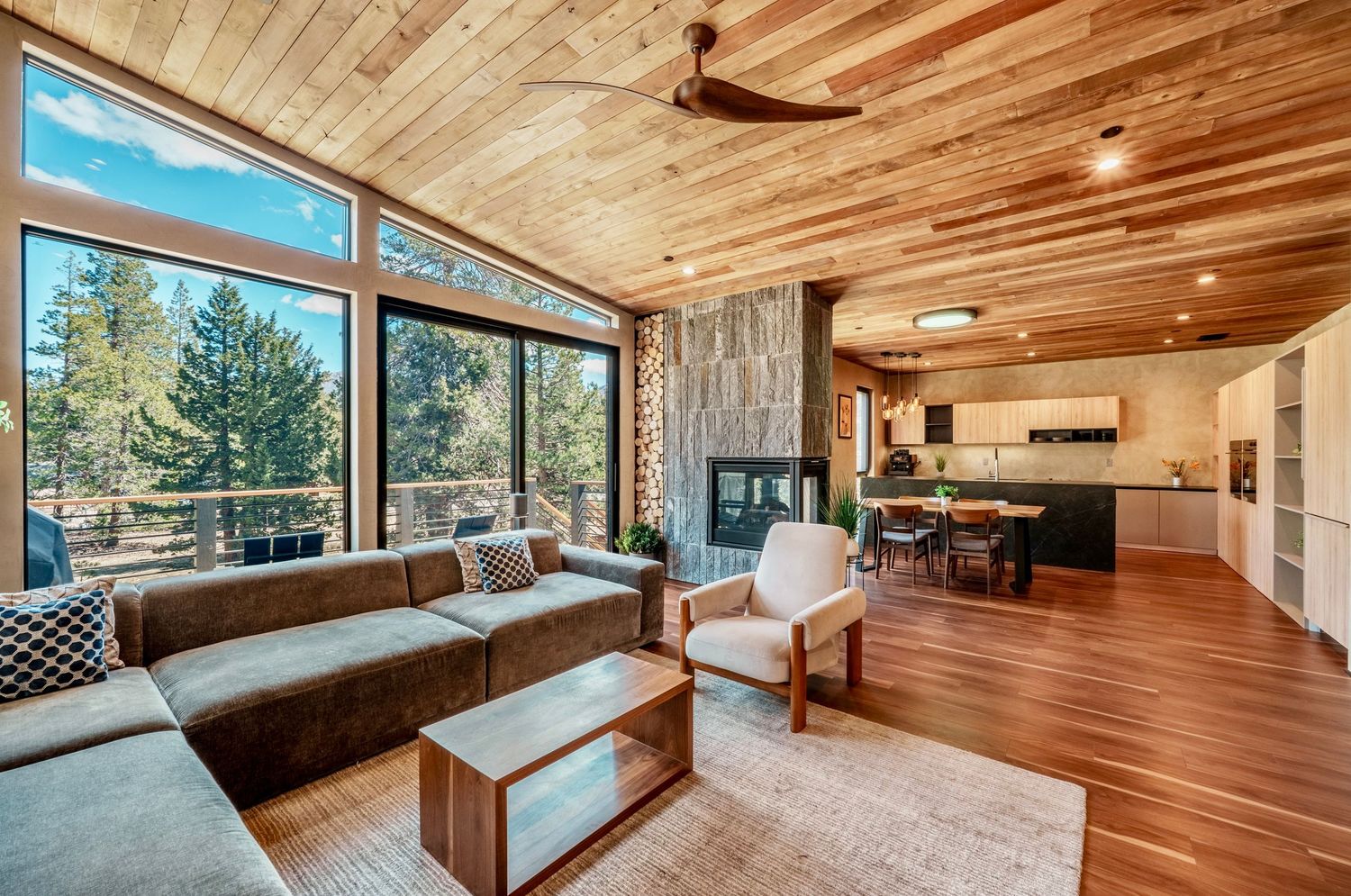 Modern open-plan living room with wood ceiling, large windows, stone fireplace, and hardwood floors in a mountain home.