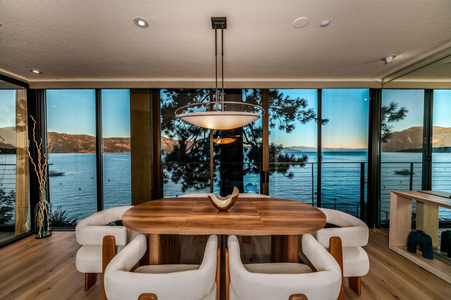 Modern dining room with walnut table, white chairs, and panoramic floor-to-ceiling windows overlooking a scenic waterfront at sunset.