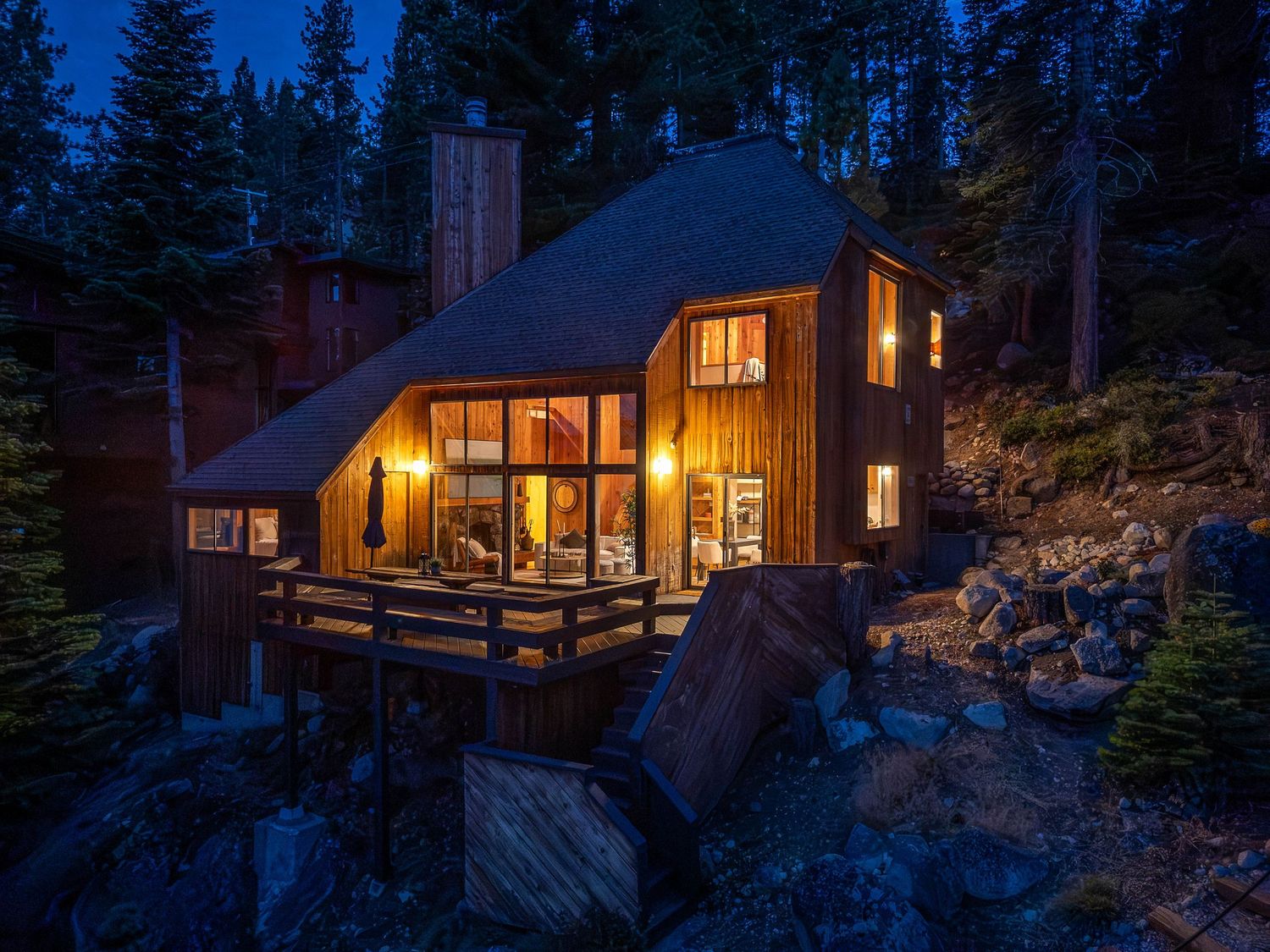 Rustic wooden chalet cabin glowing warmly at dusk, nestled among pine trees and rocky terrain in a mountain setting.