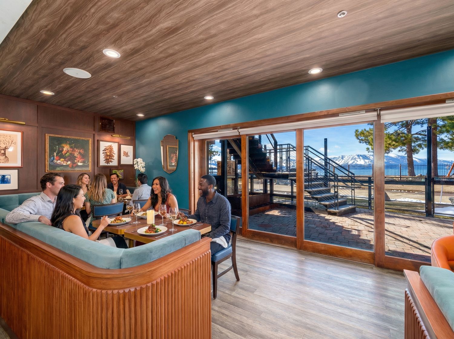 Cozy ski lodge bar with wood paneling, teal walls, and large windows overlooking snowy mountain slopes and pine trees.