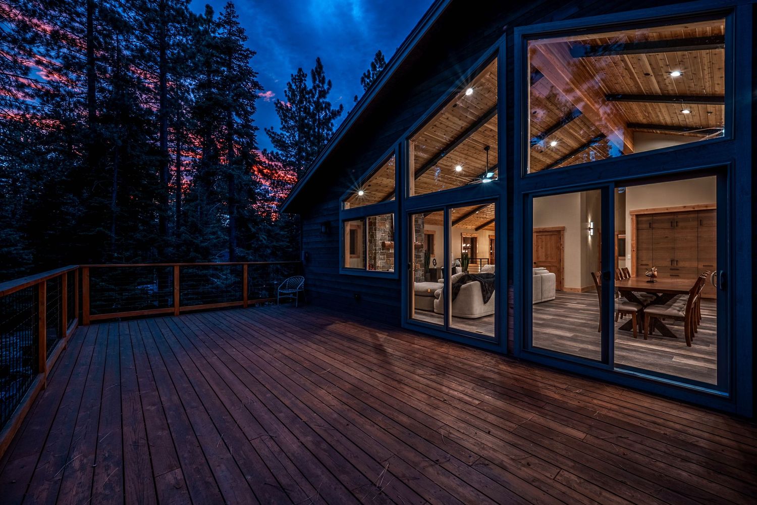 Illuminated modern cabin deck at dusk with large windows revealing warm interior lighting surrounded by tall pine trees.