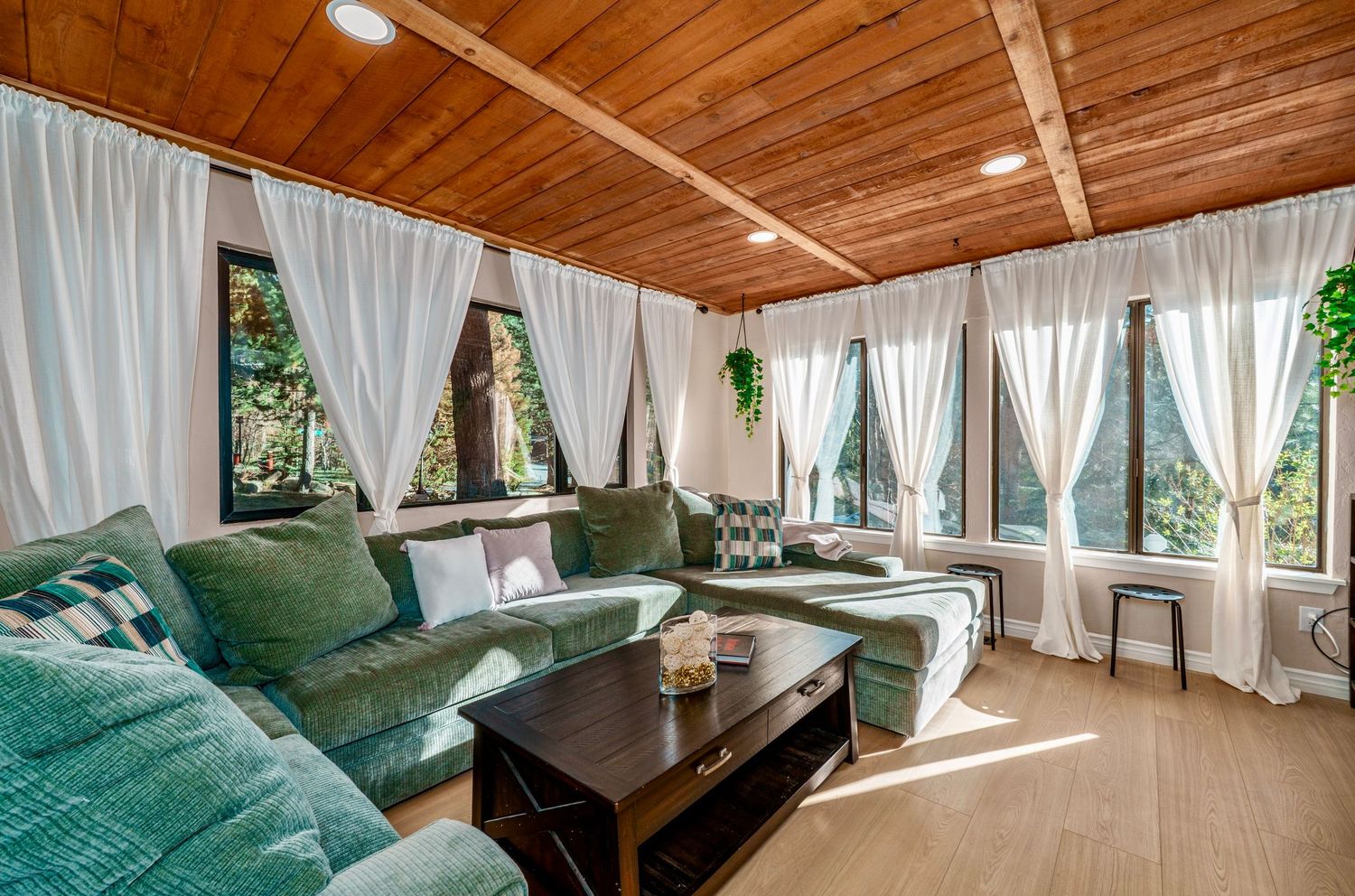 Sunlit living room with teal sectional sofa, wood plank ceiling, sheer white curtains, and large windows overlooking lush greenery.