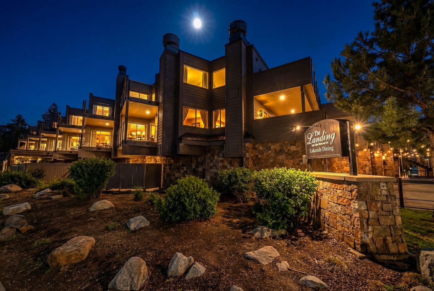 Multi-story mountain lodge with warmly lit windows and stone landscaping glowing at dusk beneath a full moon and evergreen trees.