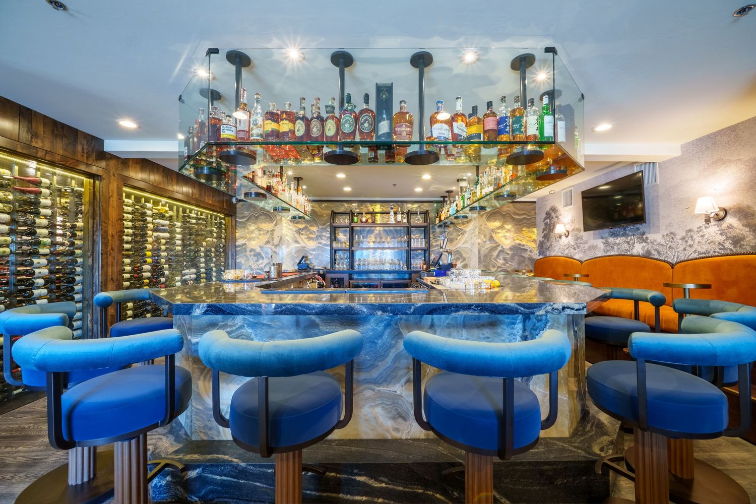 Elegant bar with blue velvet stools, marble countertop, and illuminated overhead liquor display in upscale restaurant.
