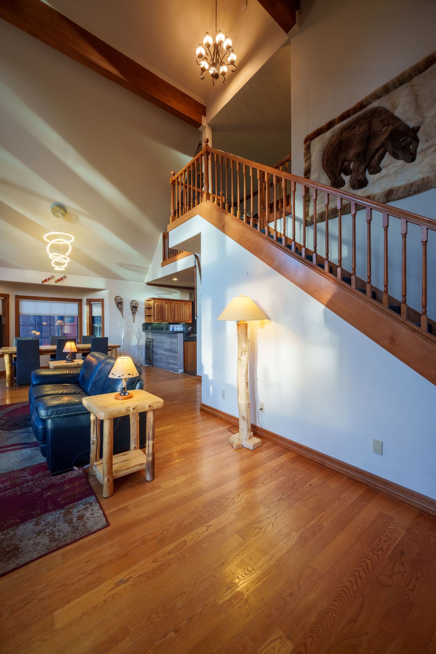 Rustic lodge living room with hardwood floors, vaulted ceilings, wooden staircase, chandelier, log lamp, leather sofa and bear artwork.