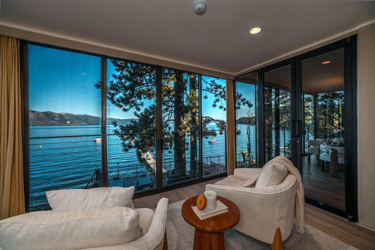 Luxury lakeside living room with floor-to-ceiling windows overlooking serene blue water and pine trees with white chairs.