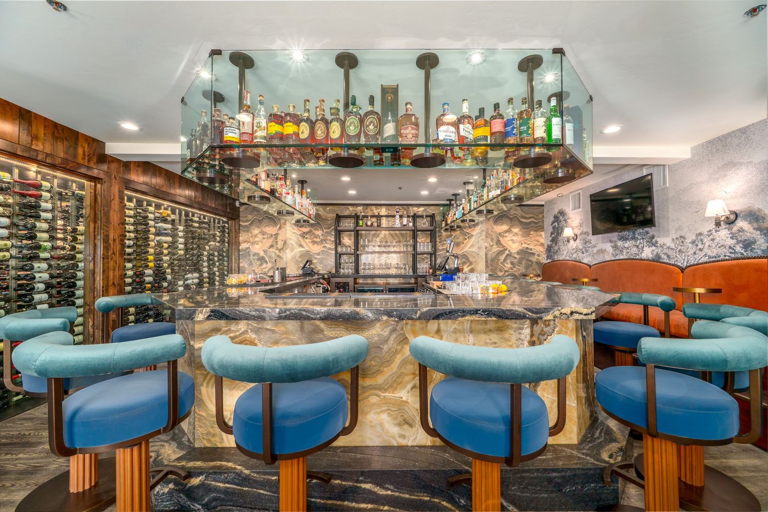 Elegant bar with blue velvet stools, marble countertop, glass liquor display, and wine wall in upscale restaurant.
