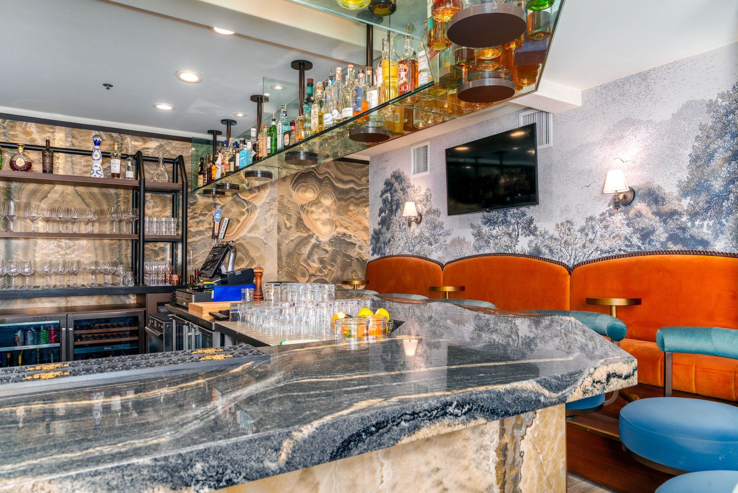 Elegant bar interior with a marble countertop, backlit bottle shelves, orange leather banquettes, blue chairs and a wall-mounted TV.