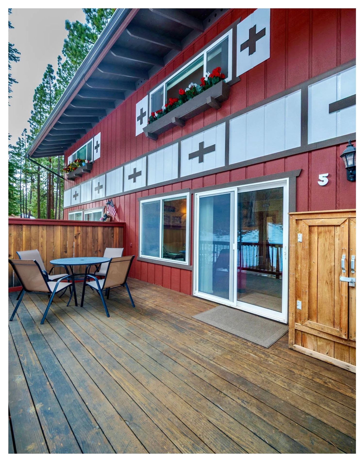 A wooden deck with railings extends from a red and white cabin with large windows and Swiss-style cross decorations.