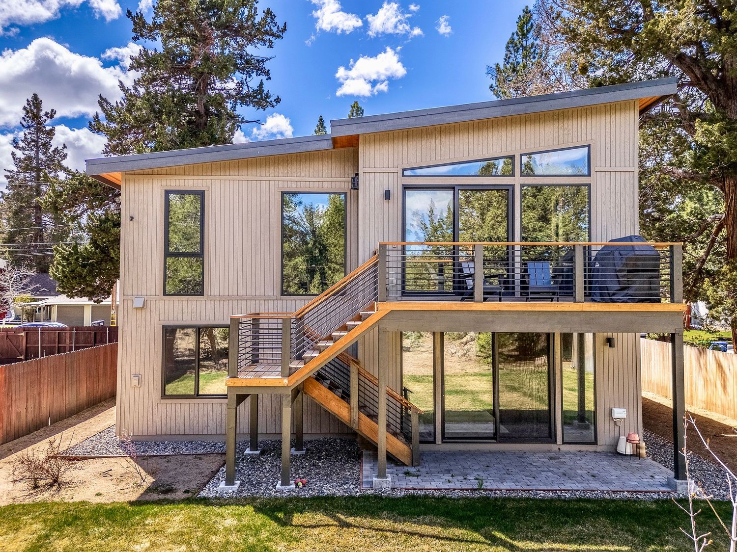 Modern two-story home with large windows, wood staircase, upper deck with cable railings, surrounded by tall pine trees.