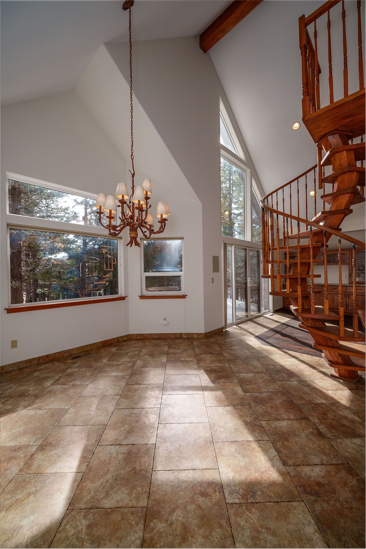 Modern living room with spiral wooden staircase, high vaulted ceiling, large windows, and elegant chandelier on brown tiled floor.