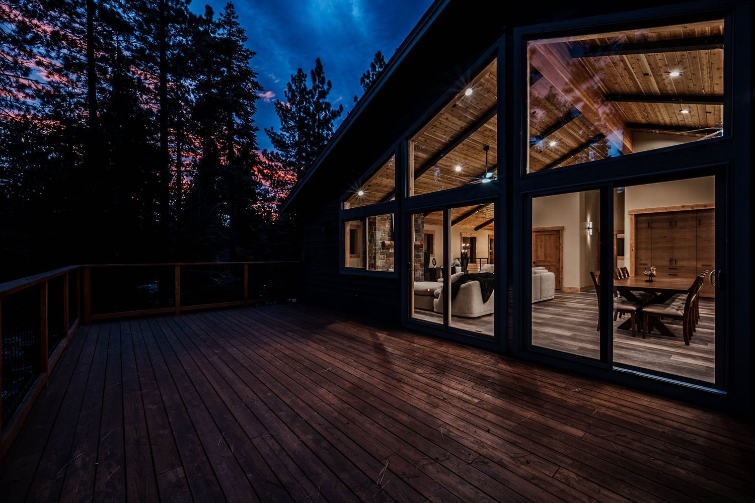 Modern cabin deck at night with warm interior lighting glowing through large glass windows surrounded by tall trees.