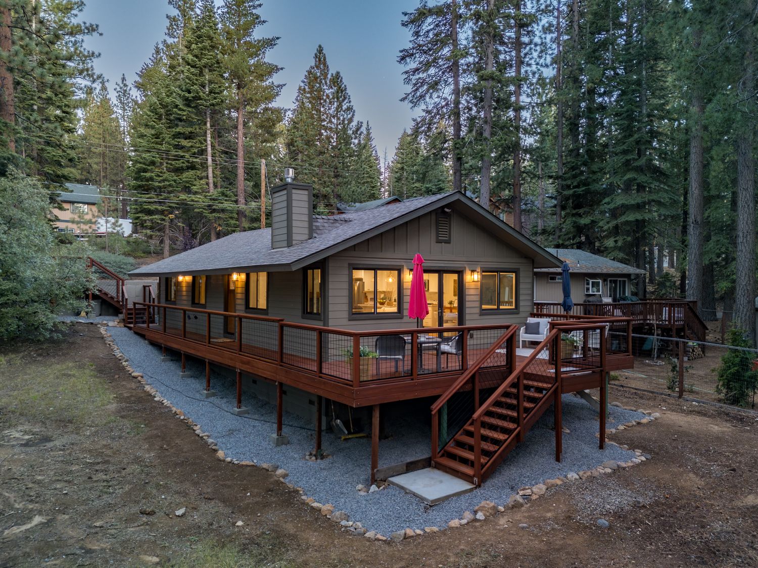 Modern cabin nestled among tall pine trees with a raised wooden deck and stone pathway in a forest setting.