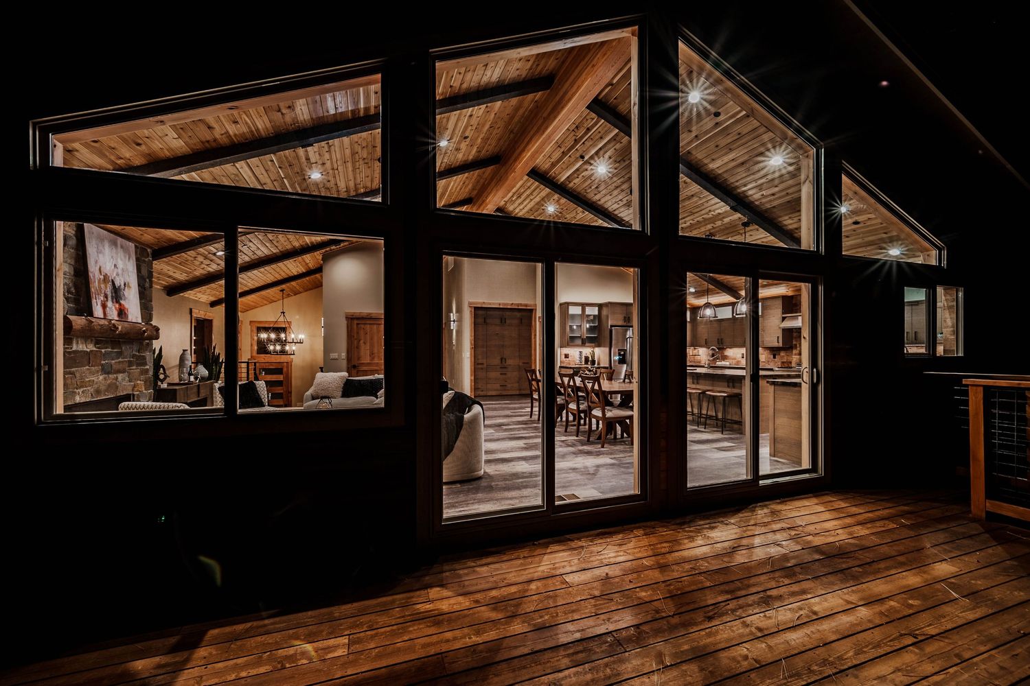 Warm wooden deck at night with large glass windows revealing a cozy lit interior of a rustic cabin home.