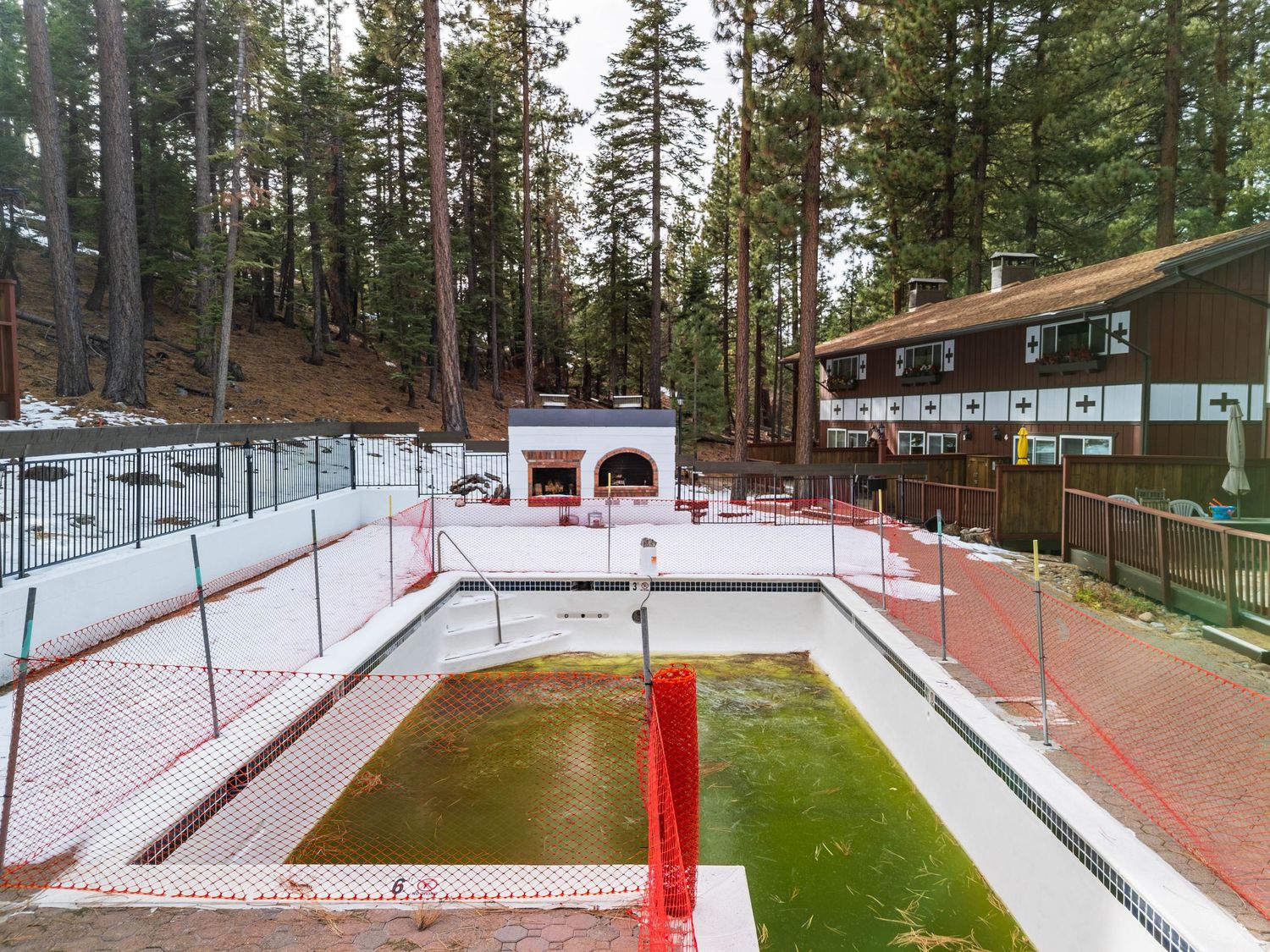 Panoramic view of a snowy ski slope with green grass patches and red poles marking the trail through pine trees.