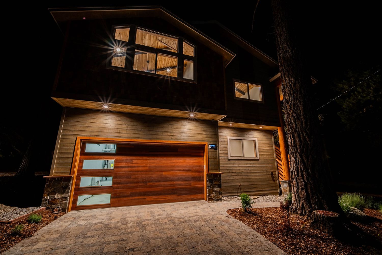 Modern two-story home at night featuring a wood-panel garage door, stone driveway, and warm exterior lighting.