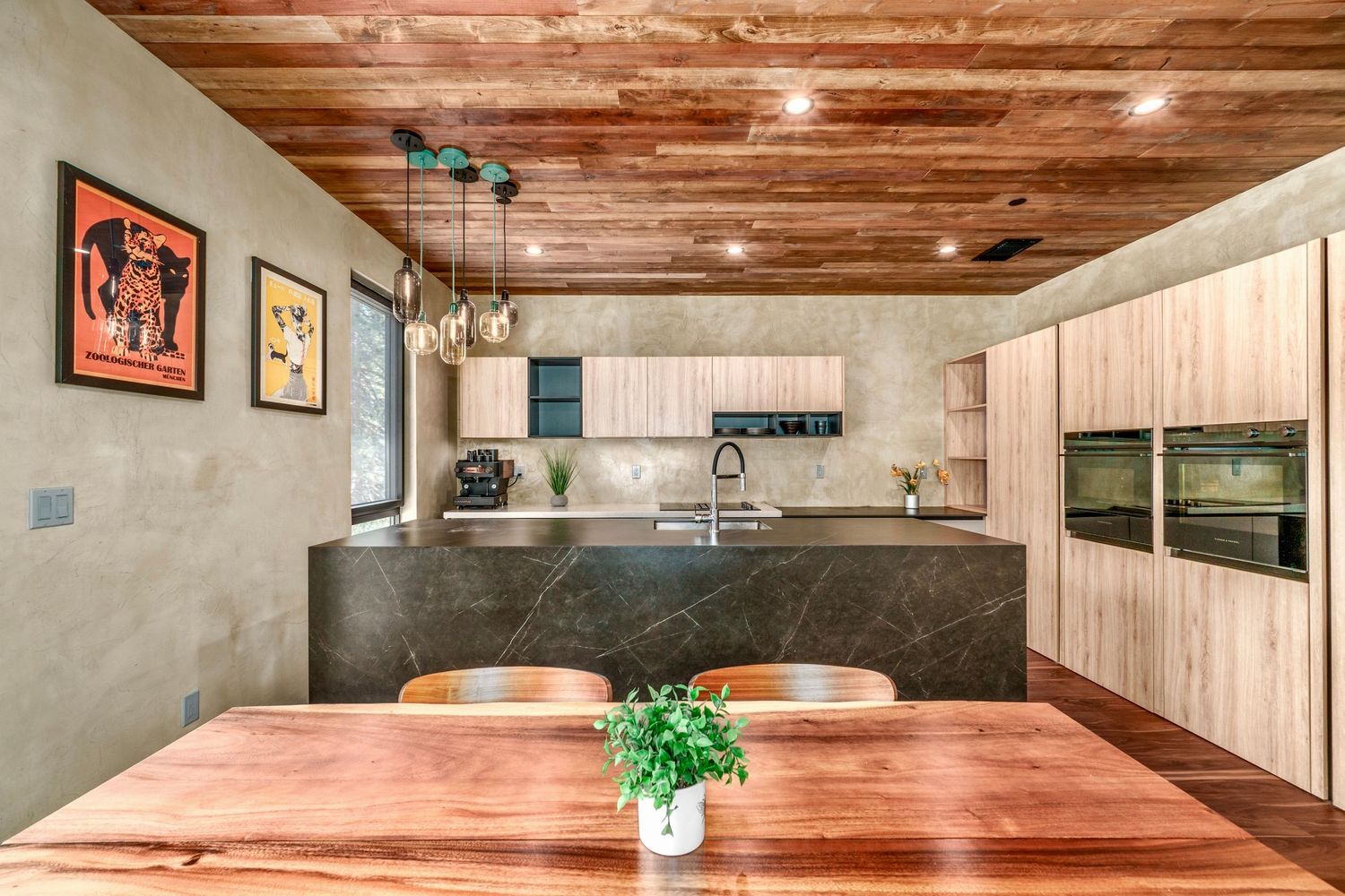 Modern kitchen with wood ceiling, marble island, light wood cabinetry, pendant lights, and a rustic dining table with chairs.
