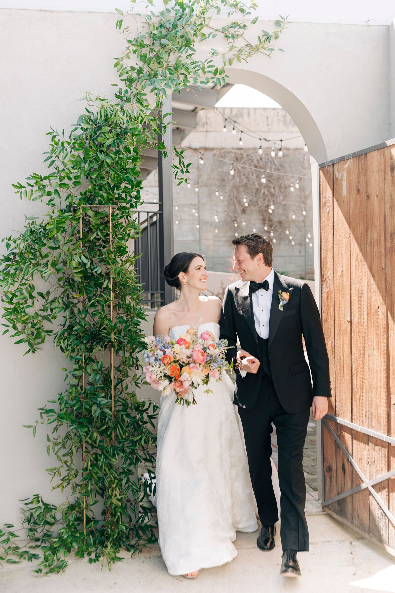 Bride and groom walk through floral arch with greenery, bride holding colorful bouquet in elegant white gown and black tuxedo.