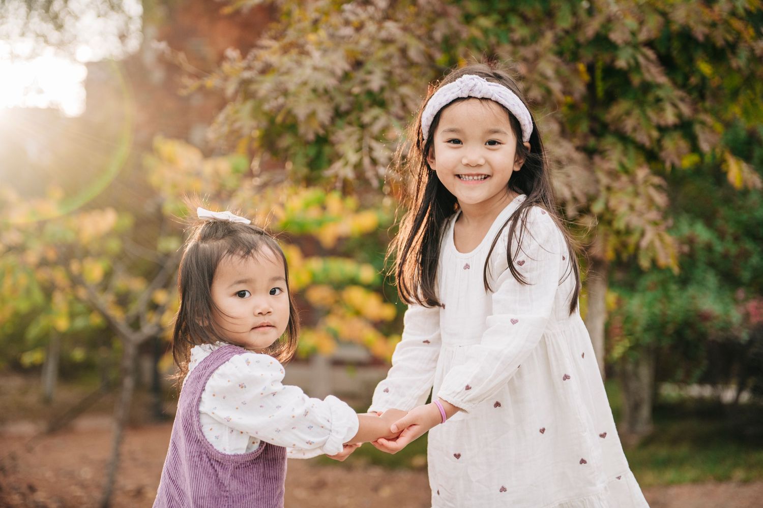 Two young Asian girls in dresses hold hands outdoors, smiling among autumn trees in warm golden light.