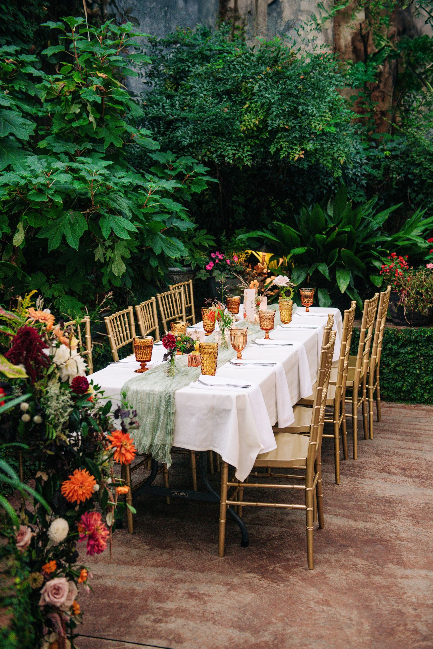 Elegant outdoor garden wedding reception table with white linen, gold chairs, colorful glassware, and lush tropical greenery backdrop.