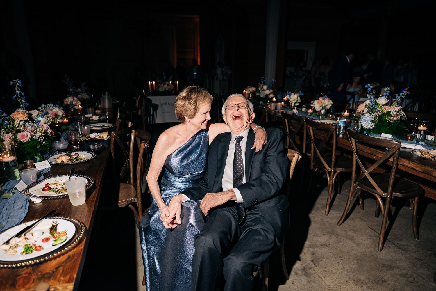 Elderly couple laughs and embraces joyfully while seated at elegantly decorated wedding reception table.