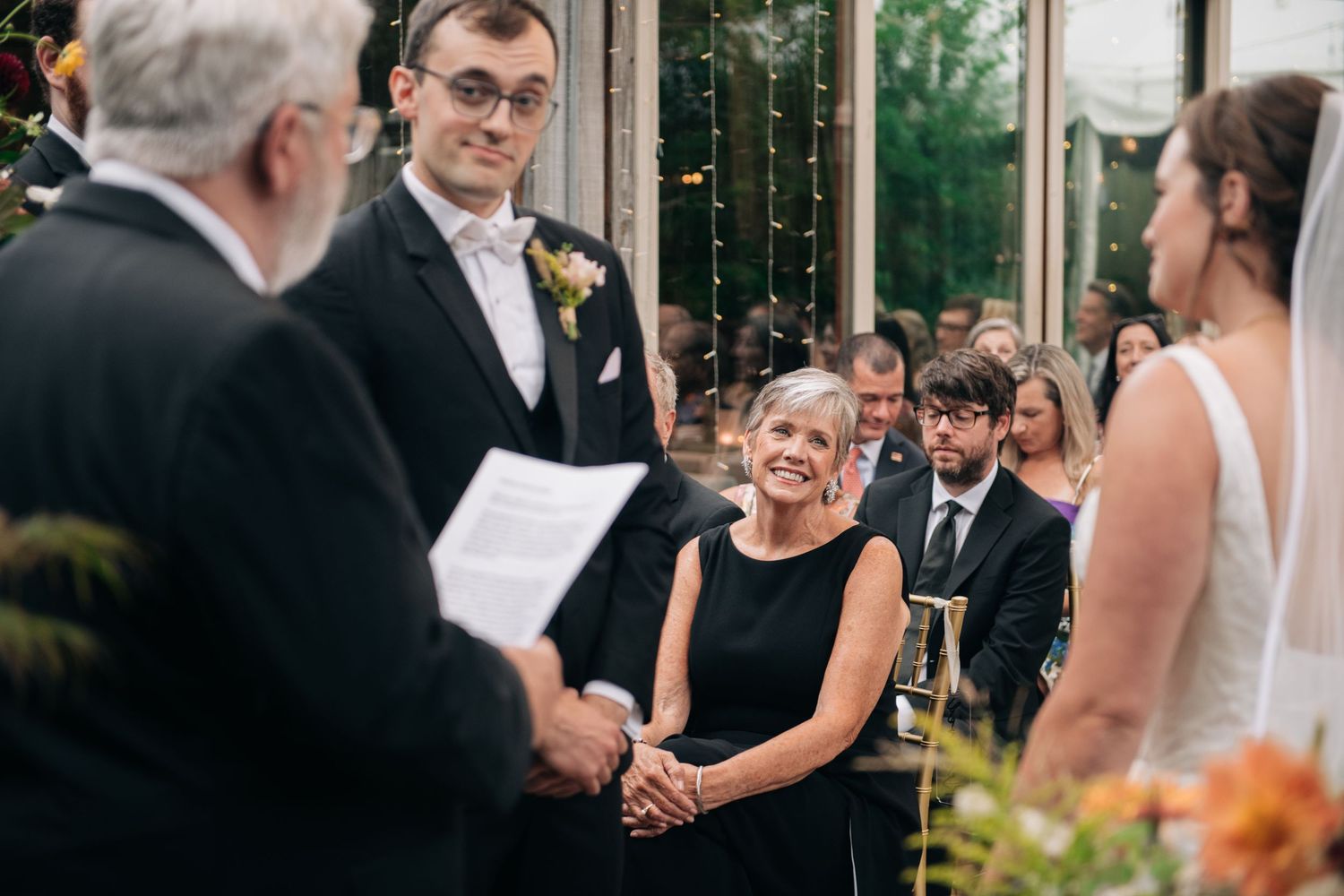 A groom in a suit smiles at a woman in black during an outdoor wedding ceremony with guests seated in the background.