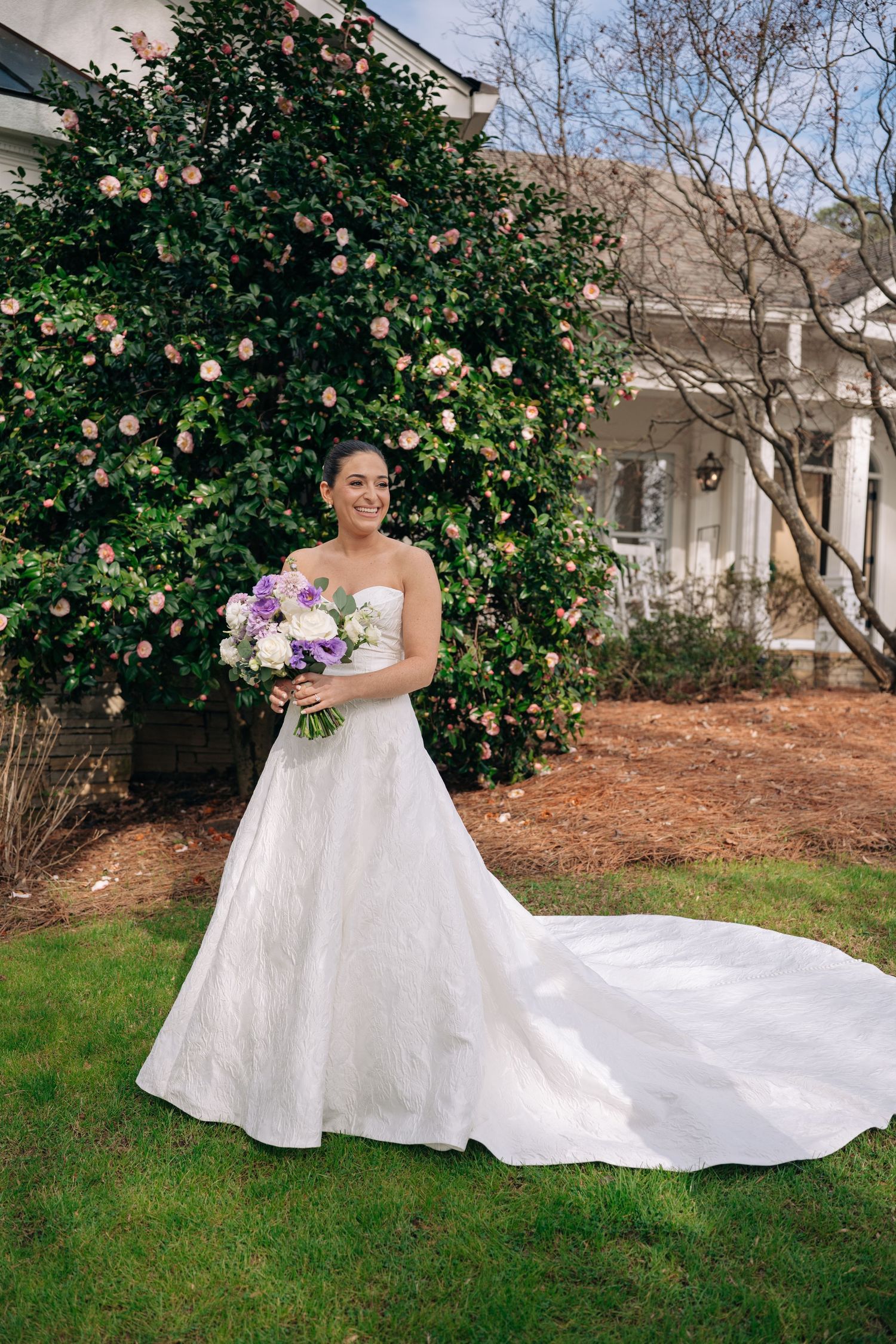 Bride in strapless white ball gown with train holds purple bouquet outdoors near flowering bush.