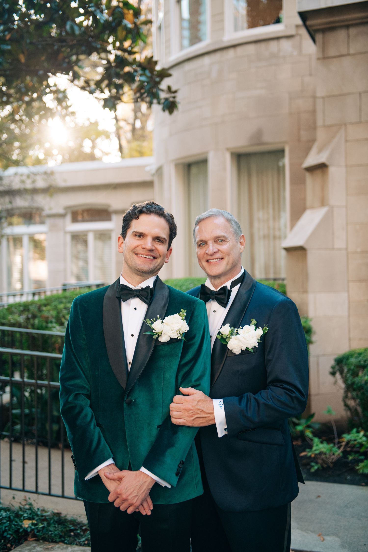 Two grooms in green and navy velvet tuxedos with white boutonnieres pose together outdoors.
