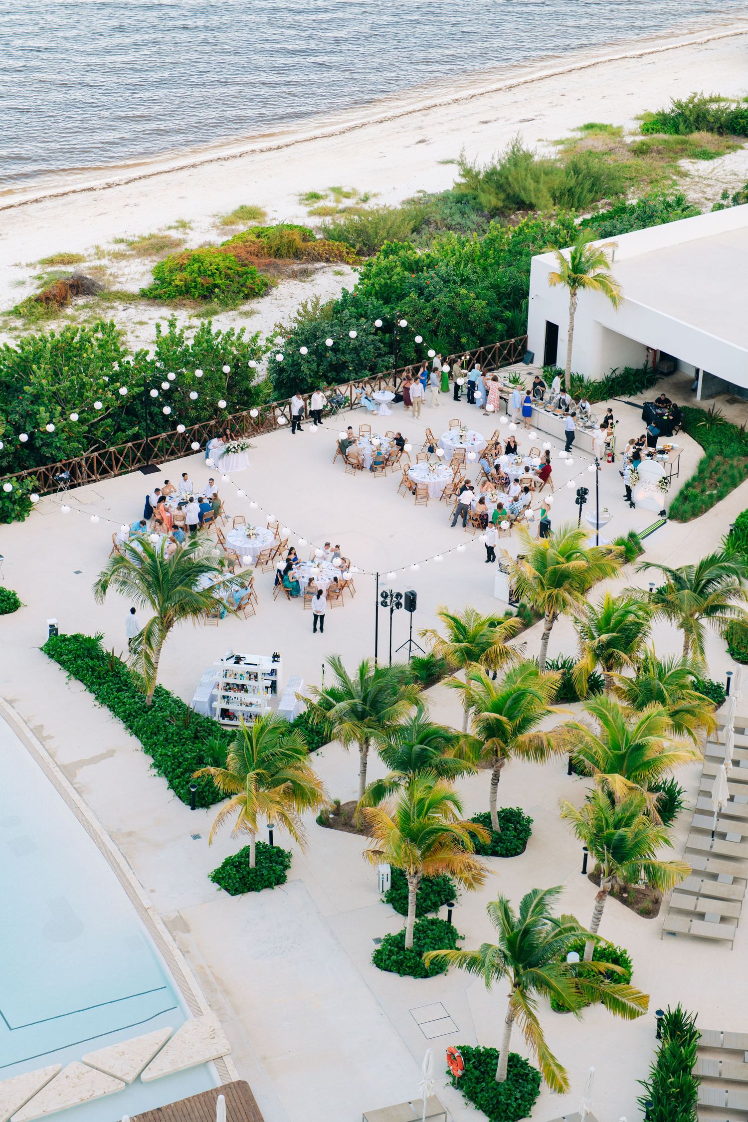 Aerial view of an elegant beachside wedding reception with round tables, palm trees, string lights, and ocean backdrop.