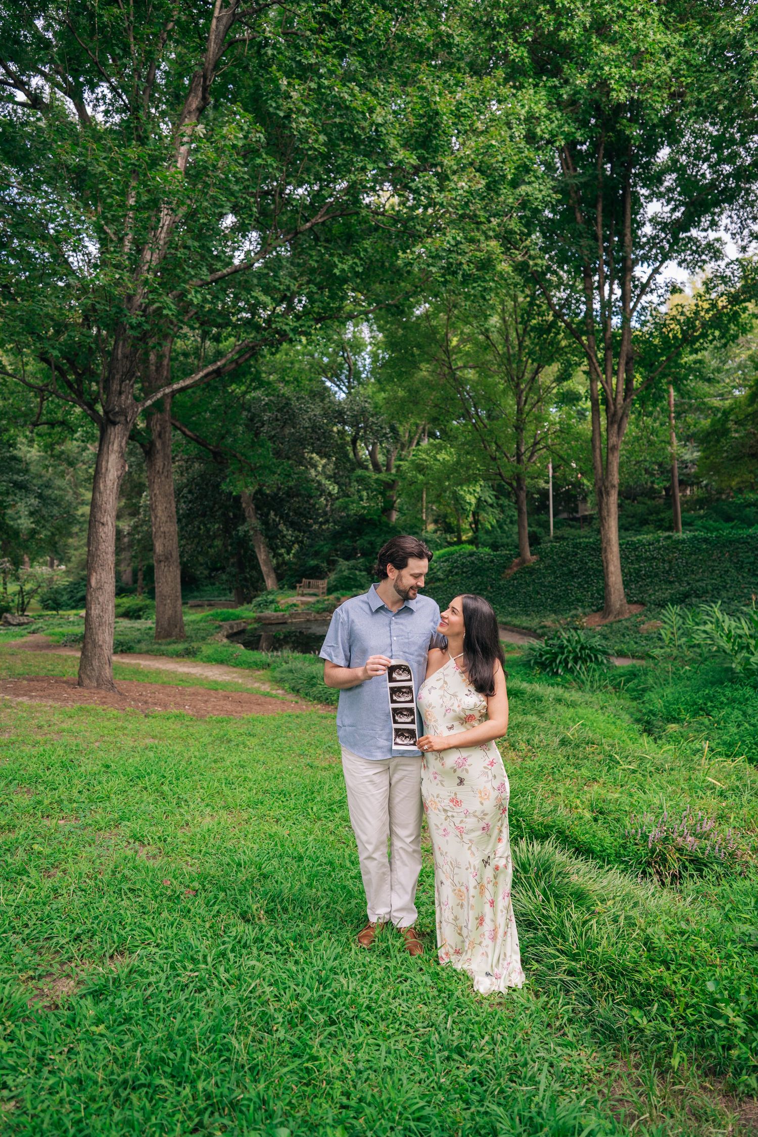 Couple holding ultrasound photos in lush green park, pregnancy announcement portrait outdoors.
