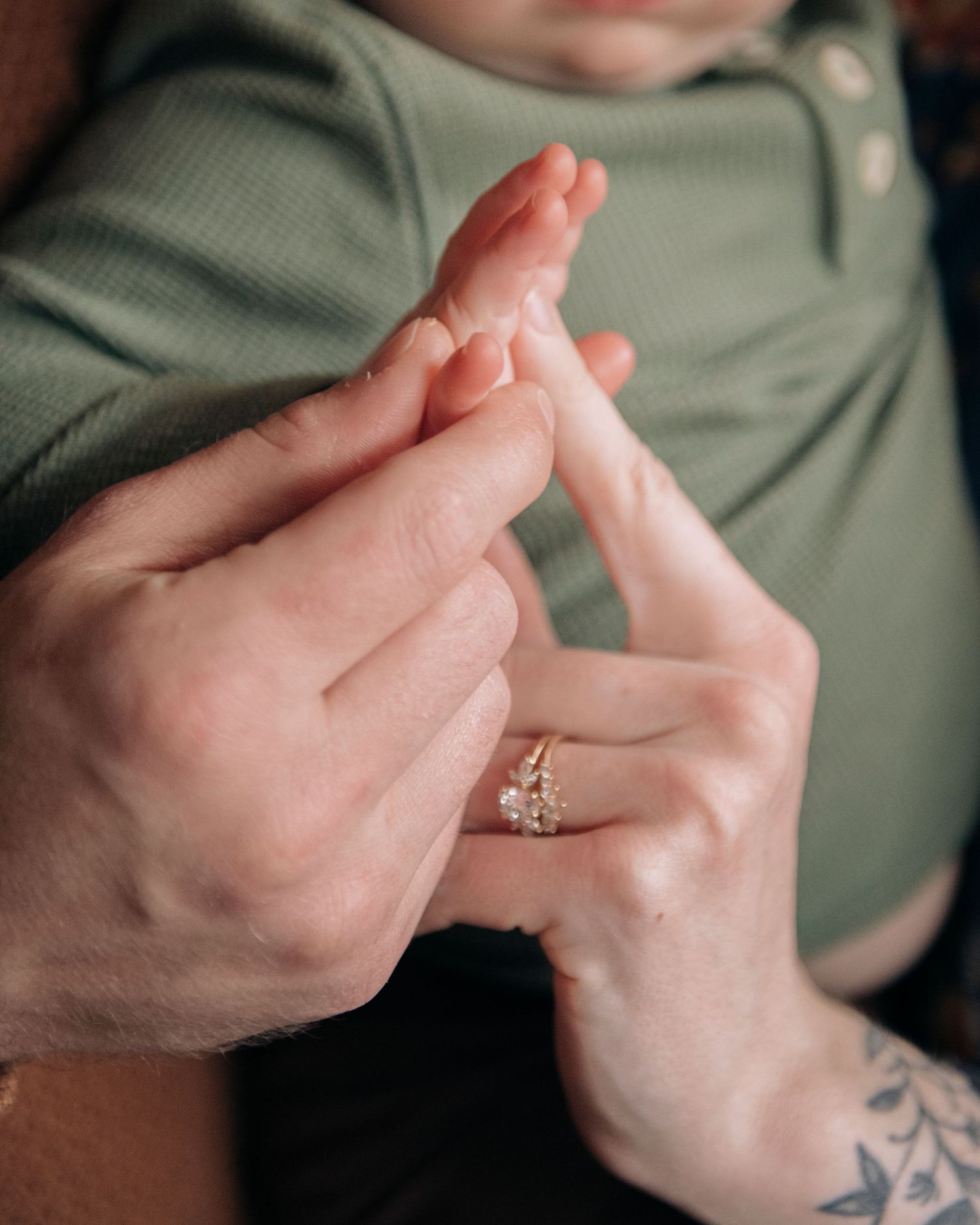 Adult hands gently holding a newborn baby's tiny feet, with a diamond engagement ring visible on one finger.