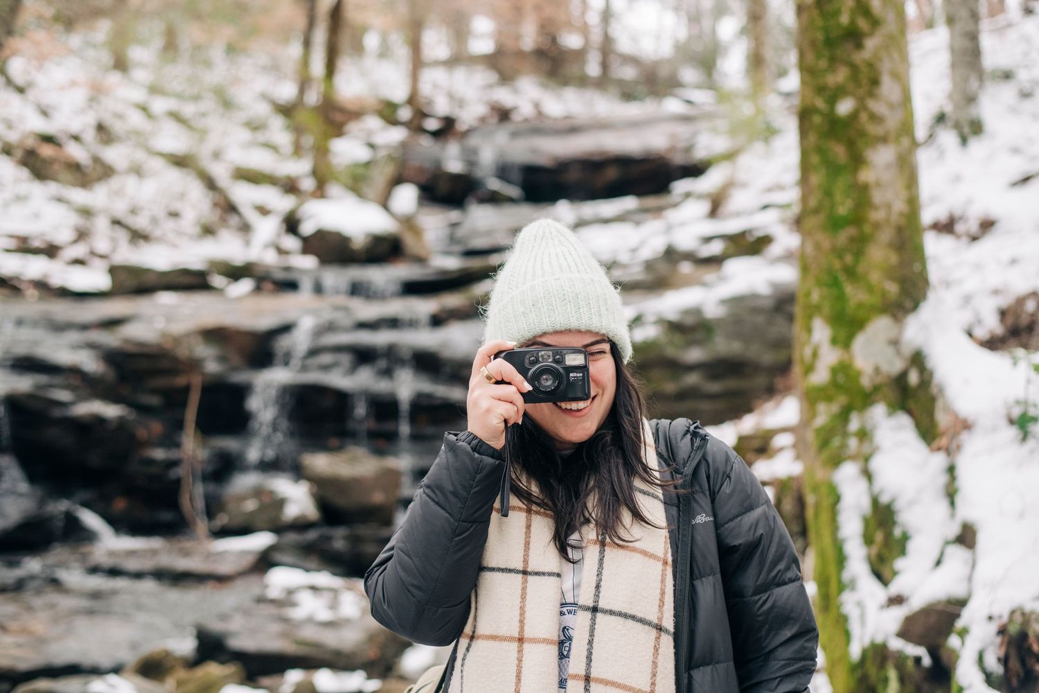 Woman in winter hat photographing with film camera near snowy waterfall in forest.