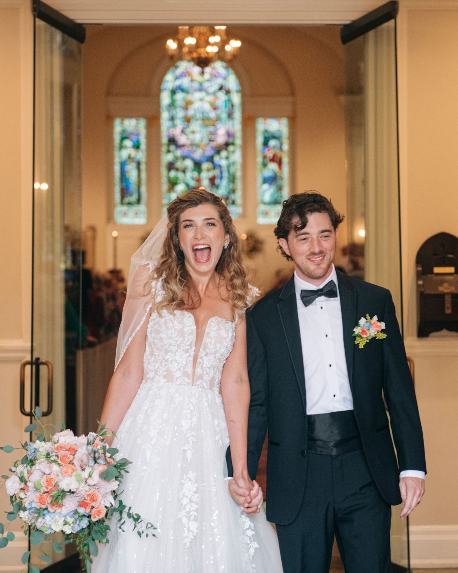 Joyful newlyweds exit church ceremony holding hands with stained glass window backdrop.