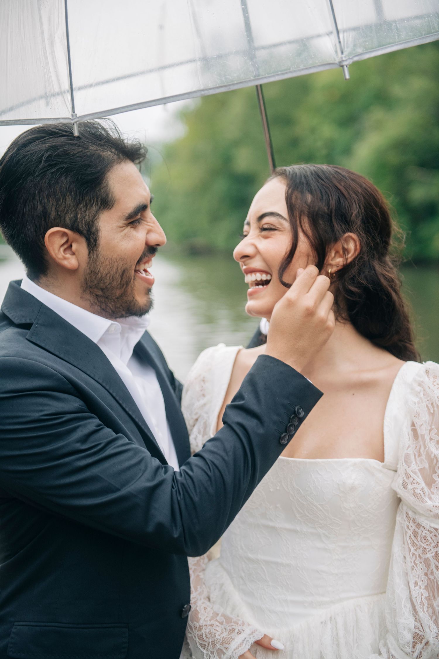 Joyful bride and groom laugh together under clear umbrella on rainy wedding day outdoors.