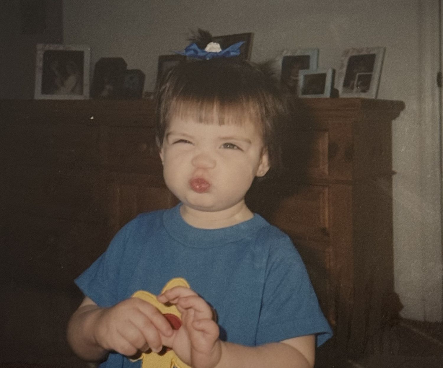 Cute toddler girl with blue bow making a pouty face, wearing a blue shirt, with picture frames on a wooden dresser behind her.