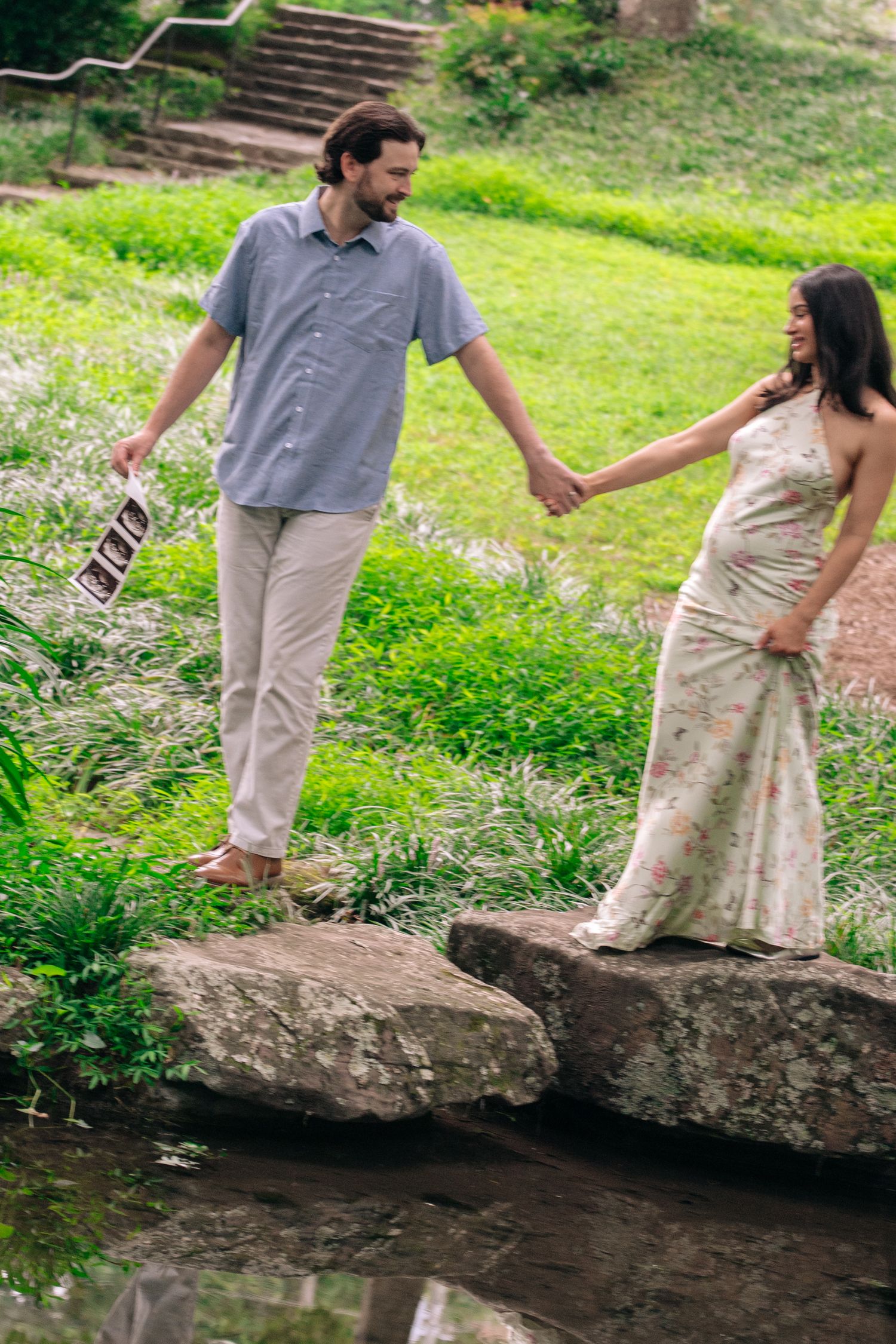 Pregnant woman in floral dress holds hands with man holding ultrasound photos while walking on stepping stones in lush park.
