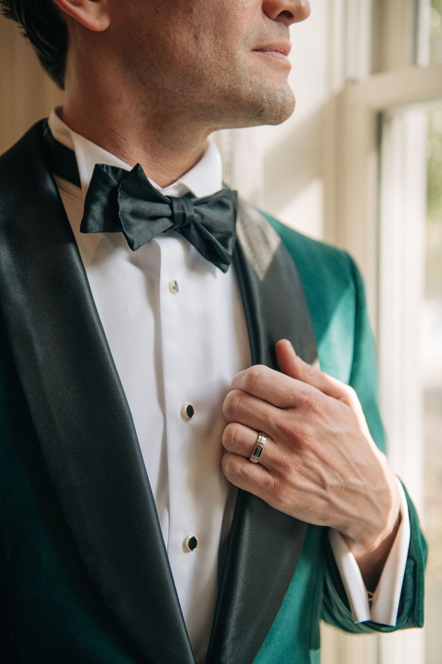 Groom in emerald green velvet tuxedo with black bow tie adjusts jacket near window light.