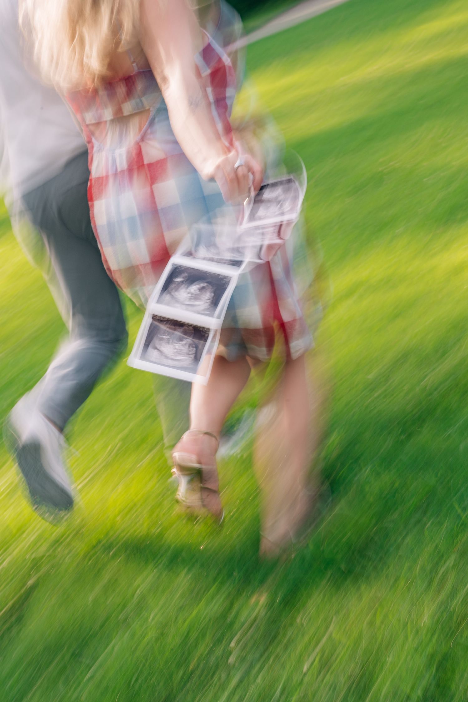 Blurred motion photo of a couple running on green grass, holding ultrasound photos, celebrating pregnancy news outdoors.