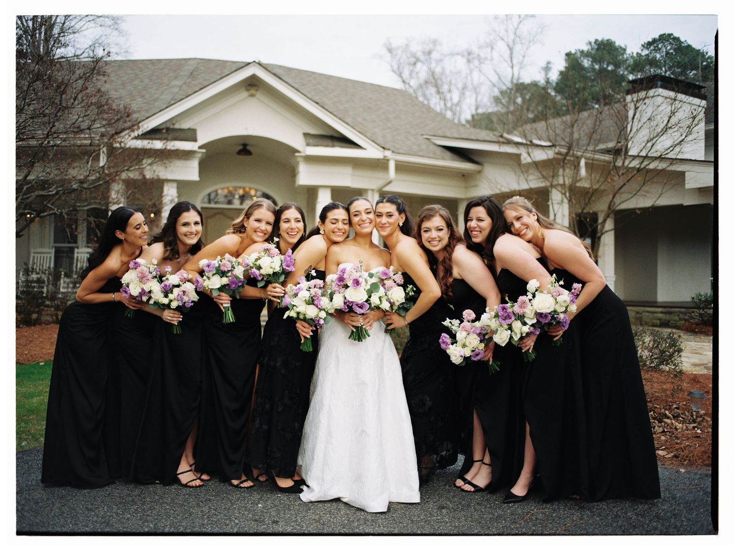 Film photo of a bride posing with bridesmaids in black dresses holding pink and white bouquets in front of Little River Farms in Georgia.