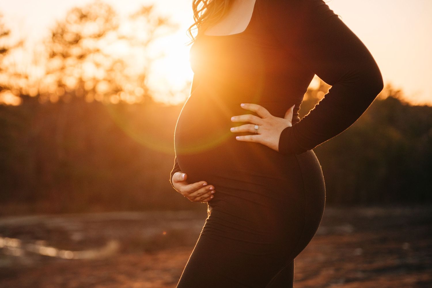 Pregnant woman in black dress cradling her baby bump at golden sunset outdoors.