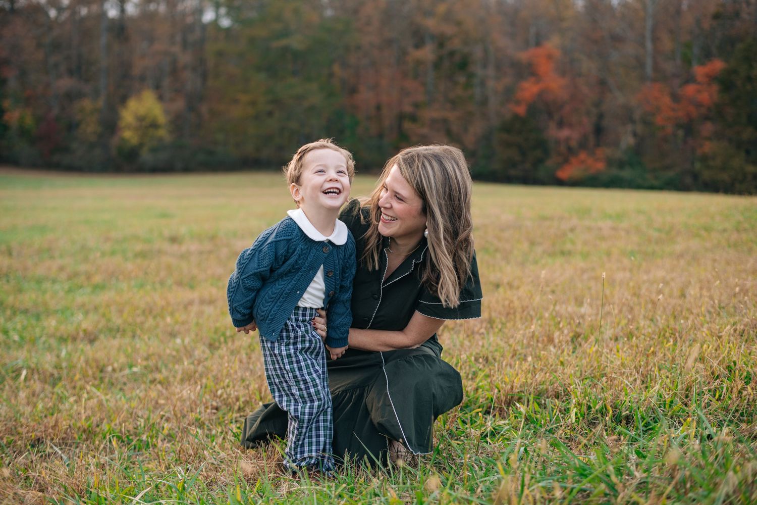 A mother and young child laughing together in an autumn field with colorful fall foliage in the background.