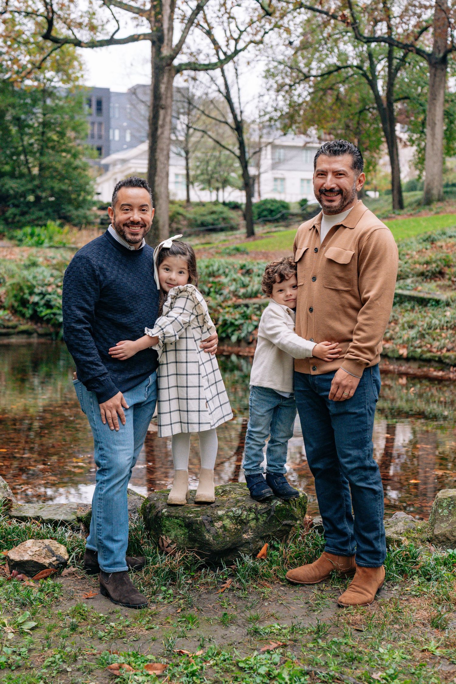 Two men smile with two young children on a rock by a pond in a lush green park during autumn.