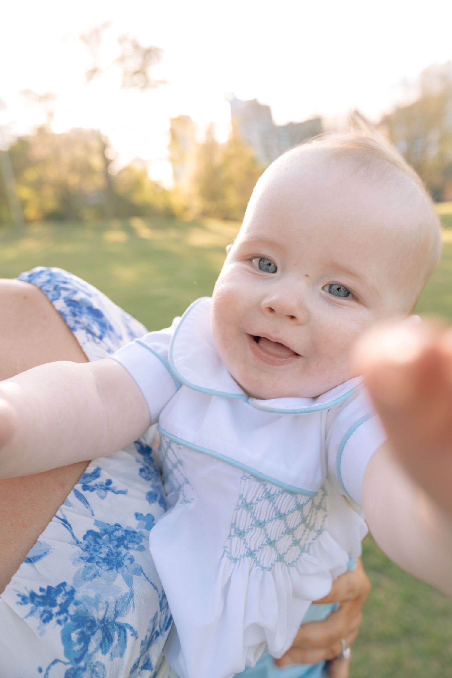 Smiling baby with blue eyes in white smocked outfit being held outdoors in golden sunlight.