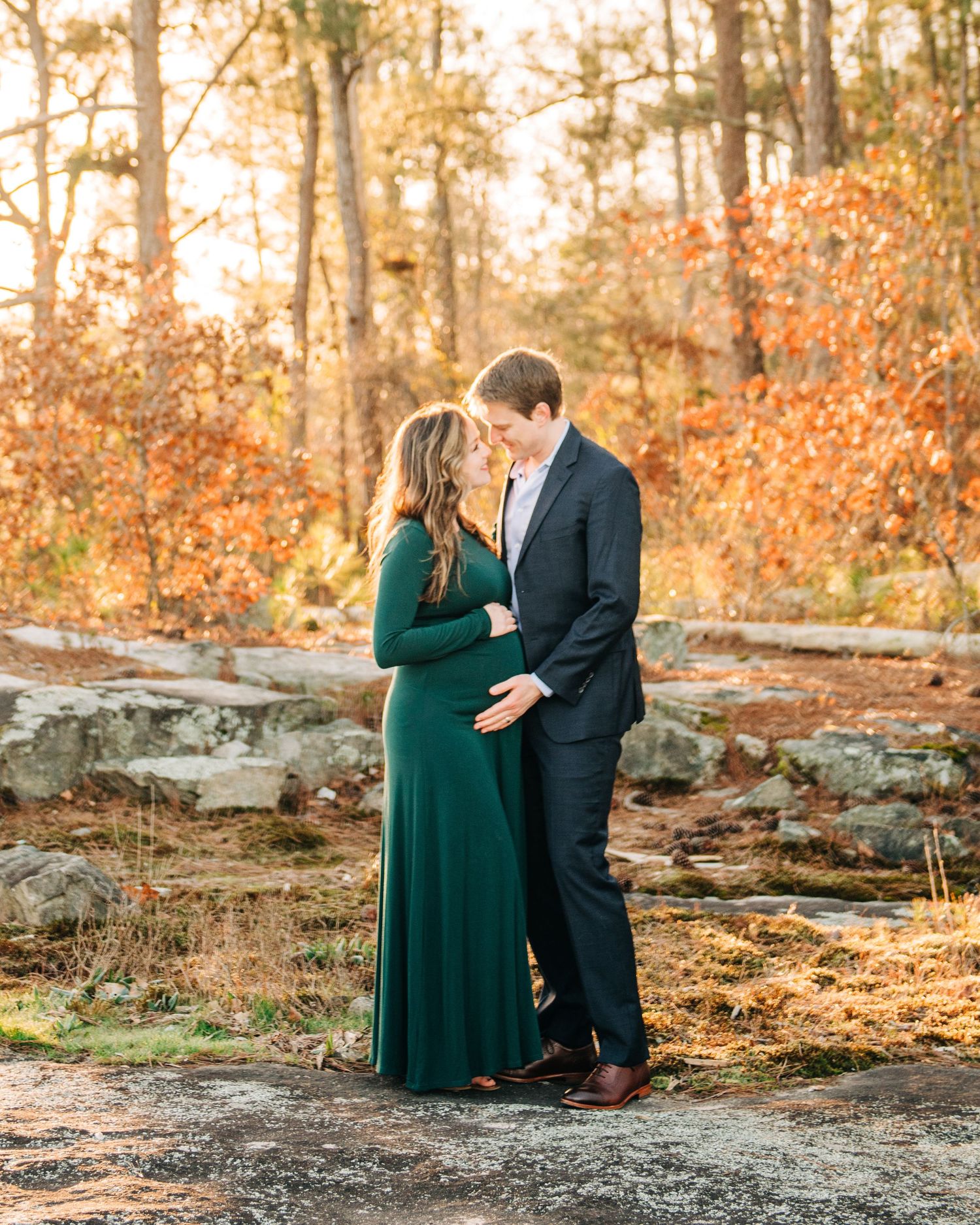 Couple sharing a tender moment outdoors in fall foliage, woman in green maternity gown, man in dark suit.