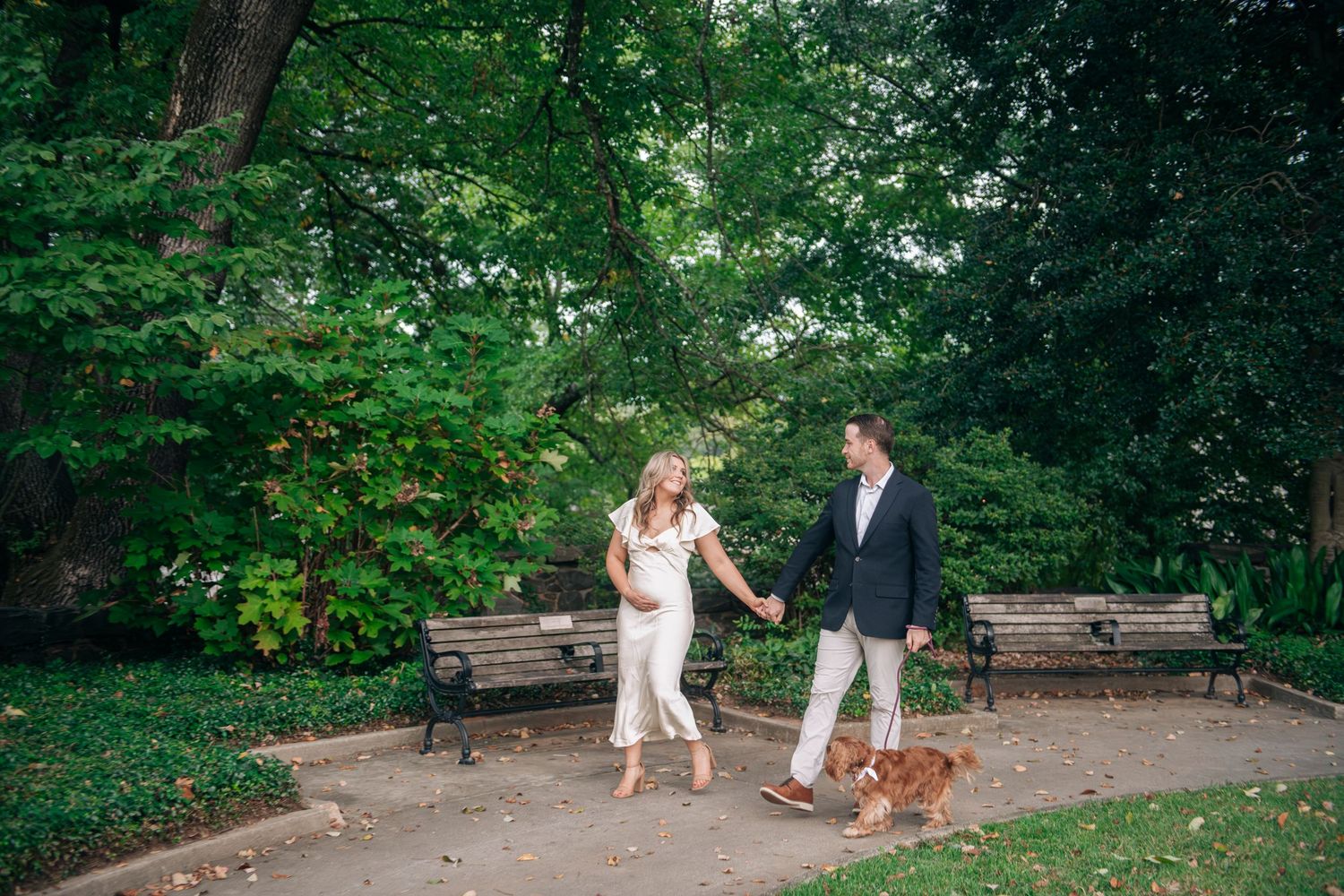 Couple holding hands walking a golden retriever dog through a lush green park with benches.