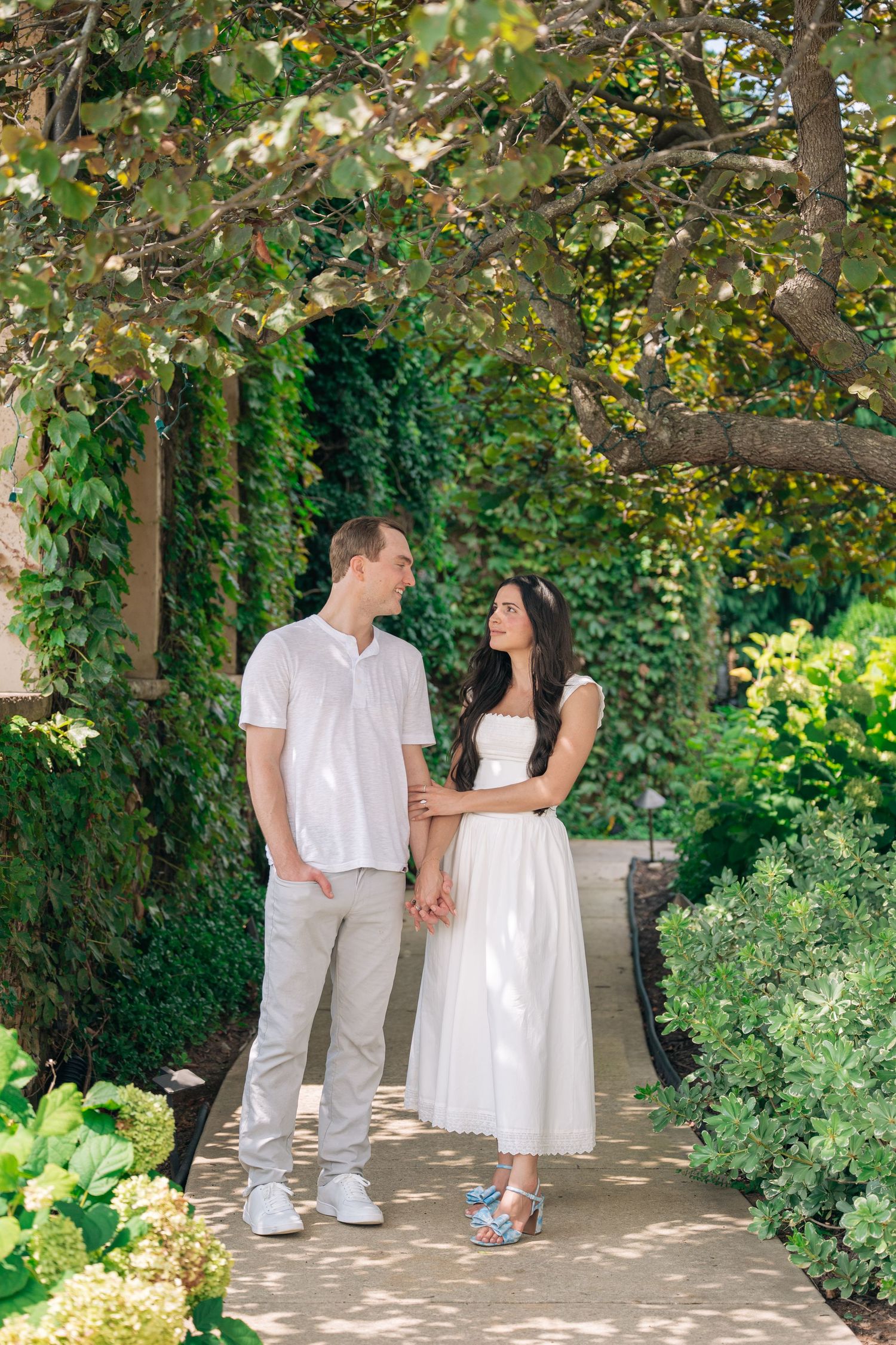 A couple in white outfits hold hands and smile at each other on a garden path surrounded by lush greenery.