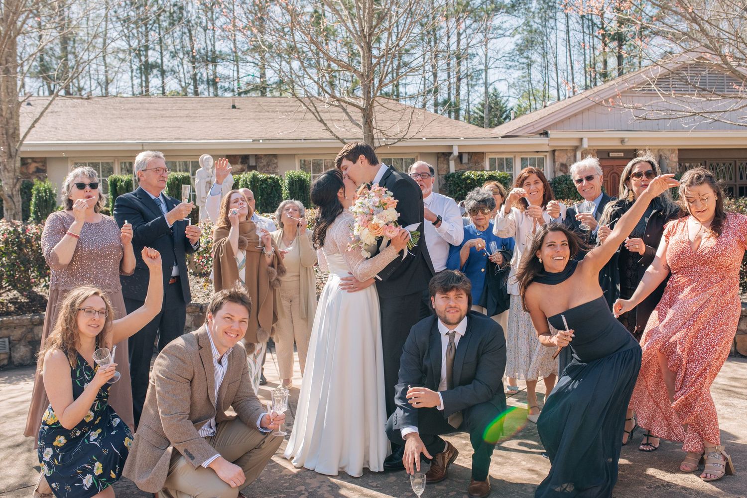 Wedding party celebrating outdoors with kissing couple surrounded by cheering guests in spring.