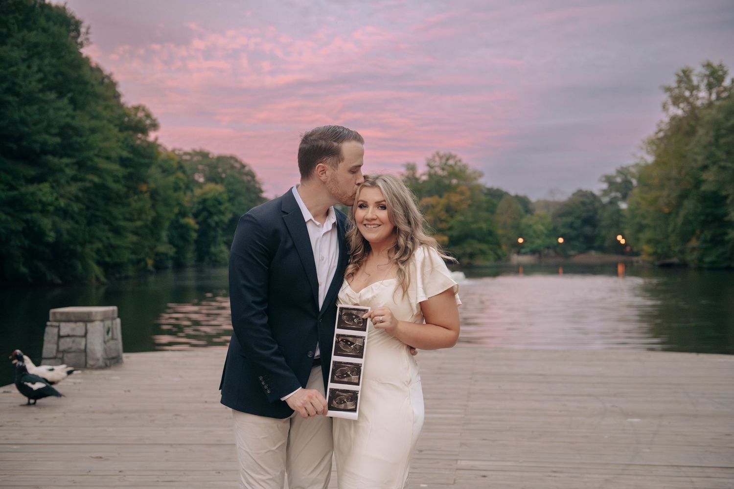 Couple holding ultrasound photos on a waterfront dock at sunset, announcing their pregnancy.