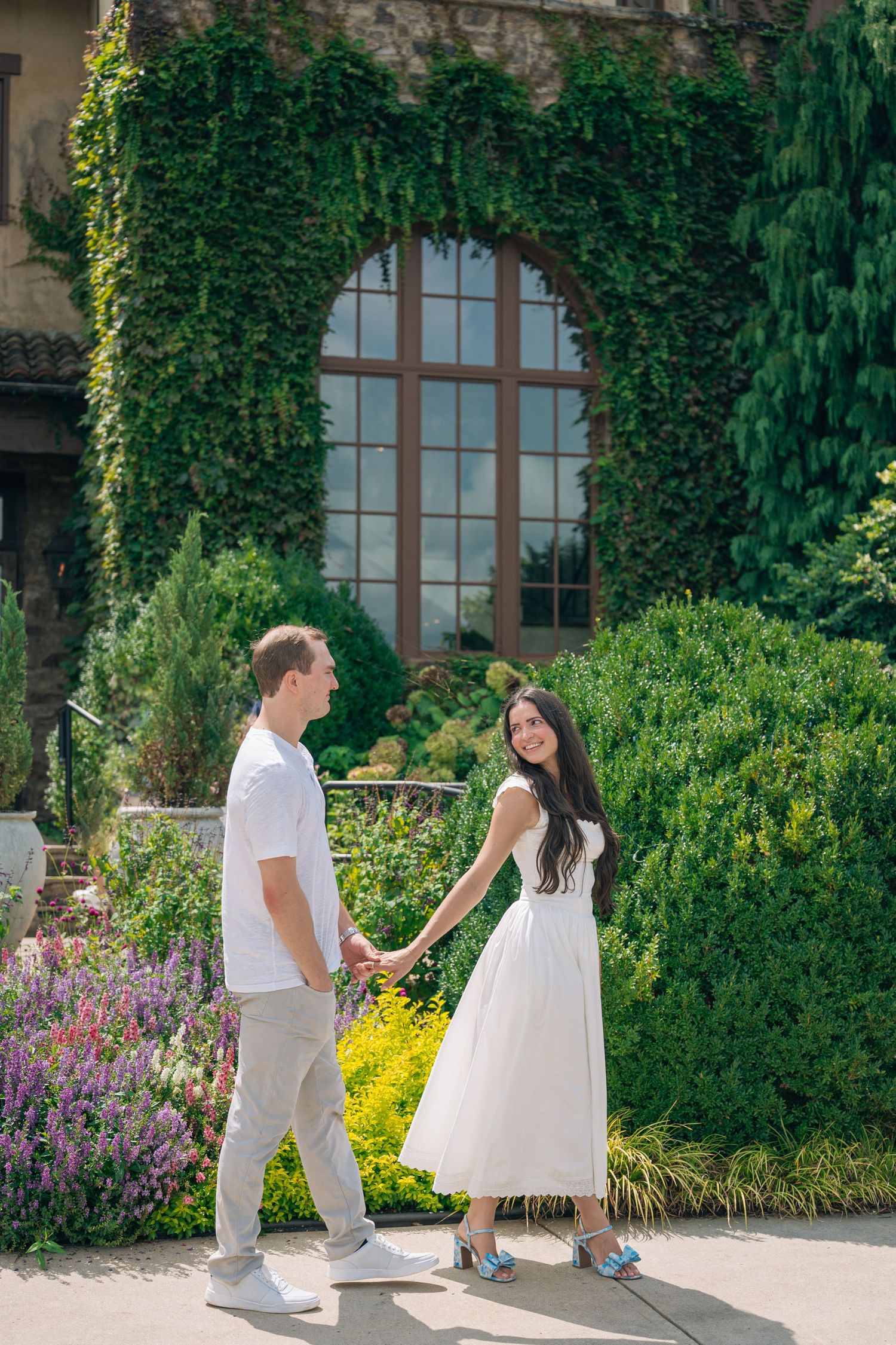 Couple holding hands in a lush garden, woman in white dress, man in white shirt, ivy-covered building backdrop.
