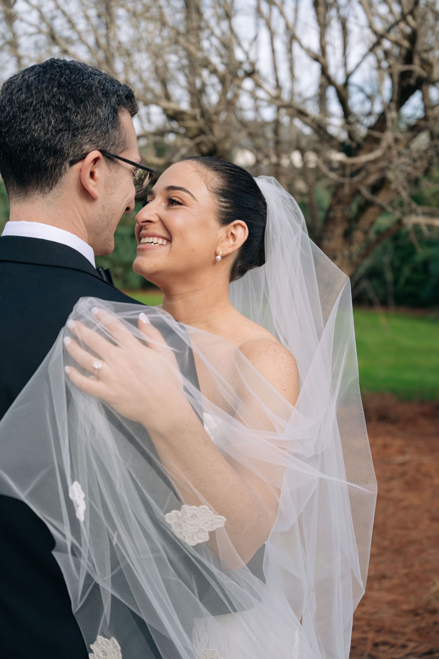 Bride in white veil smiles lovingly at groom in black suit outdoors during romantic wedding portrait.